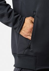 Hand inserted into the zippered pocket of a black jacket with ribbed waistband, showing a close-up of the sleeve and pocket detail.