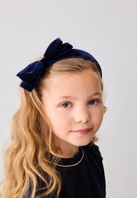 Fascia in velluto blu navy con un grande fiocco nella parte superiore e un accento di bottone dorato. Il modello ha i capelli biondi ondulati.