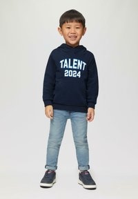 Marineblauwe hoodie met "TALENT 2024" in witte en lichtblauwe tekst, gecombineerd met lichtblauwe jeans en donkere sneakers. Soepele stof, casual pasvorm.