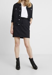 Schwarze Jeansjacke mit silbernen Knöpfen, weißes V-Ausschnitt-Shirt, schwarzer Rock mit Kordelzug und schwarze Stiefeletten. Schlichtes und modernes Design.