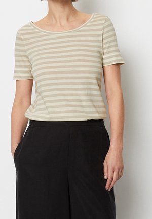 Vrouw draagt een beige-wit horizontaal gestreept kortärmelig topje, ingestopt in een zwarte broek met hoge taille, handen in de zakken.