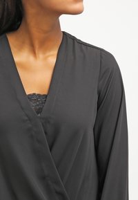 Top nero in stile incrociato realizzato in tessuto liscio, con scollo a V profondo e maniche lunghe, abbinato a una canotta con rifiniture in pizzo sottostante.