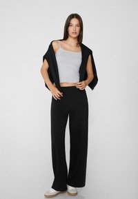 Grauer Crop-Top, schwarze weit geschnittene Hosen und weiße Sneaker. Das Model hält einen schwarzen Pullover über den Schultern. Glatte Texturen und neutrale Farben.