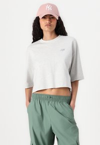 Svetlo siva skrajšana majica s kratkimi rokavi, ki ima majhen logotip New Balance, v kombinaciji s svetlo zelenimi hlačami ohlapnega kroja. Roza bejzbolska kapa.