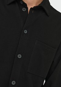 Black button-up shirt featuring a spread collar, front pocket, and textured fabric with six buttons along the placket.