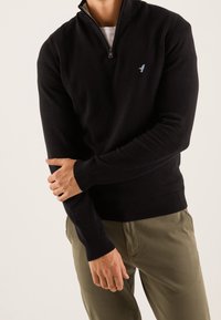 Černý svetr na zip z měkkého materiálu, s vysokým límcem a žebrovanými manžetami. Na hrudi světle modré logo ptáka.