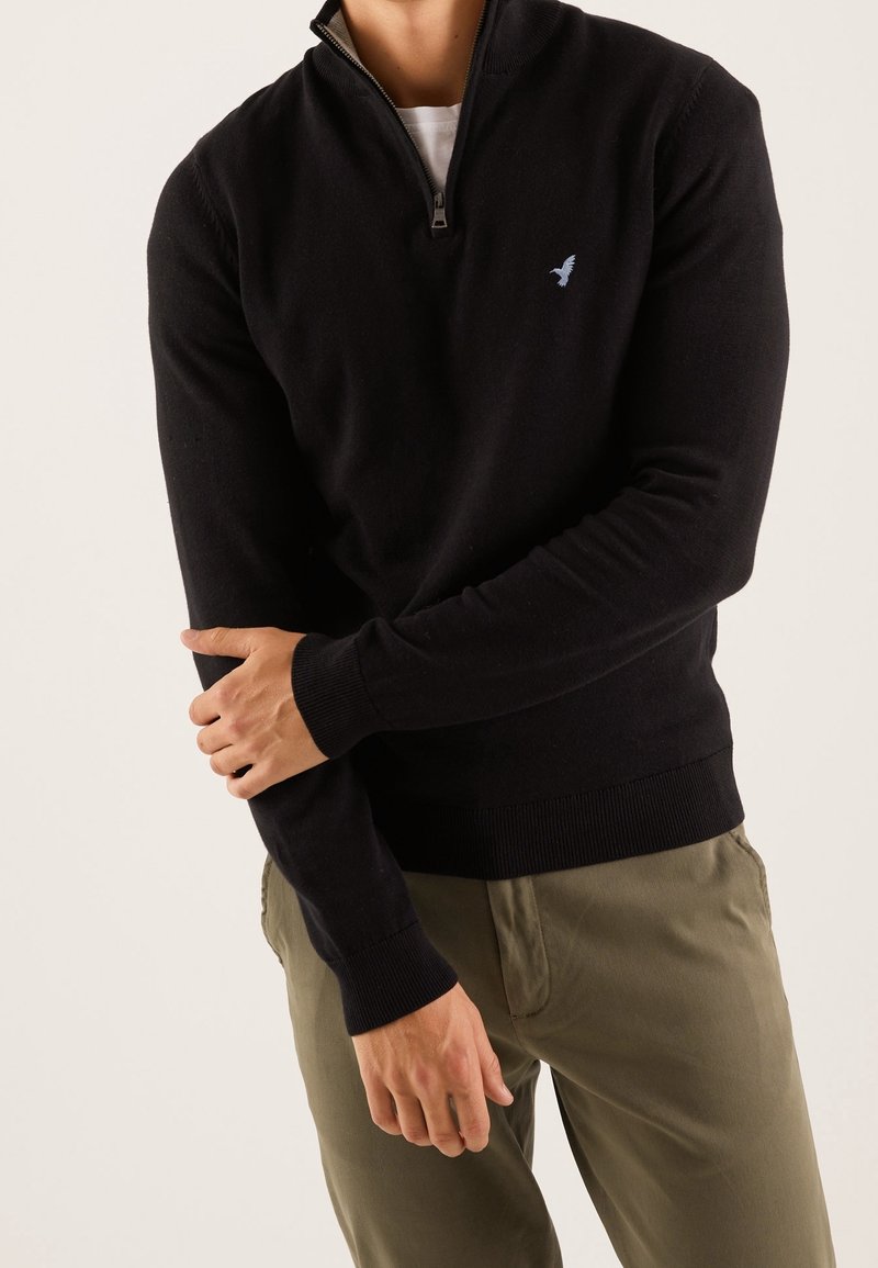 Černý svetr na zip z měkkého materiálu, s vysokým límcem a žebrovanými manžetami. Na hrudi světle modré logo ptáka.