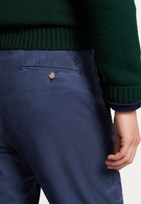 Laivansinisiä chinous-housuja, joissa on sileä pinta, takatasku nappisulkeisella ja brodeerattu logo taskun yläpuolella.