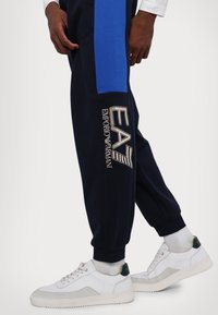 Marinblå joggers med blå sidopaneler, med en beige logotyp: "EA7 EMPORIO ARMANI." Matchas med vita och grå sneakers. Slät textur.