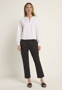 Weißes Baumwollblusenshirt mit langen Ärmeln und V-Ausschnitt, kombiniert mit schwarzen, taillierten Hosen und schwarzen Slipper-Sandalen. Neutrale Hintergrund.