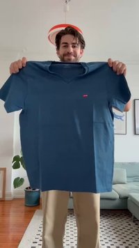 Modra bombažna majica s V-izrezom, kratkimi rokavi in rdečim logotipom Levi's na sprednji strani. Drži jo oseba v sproščenih hlačah.