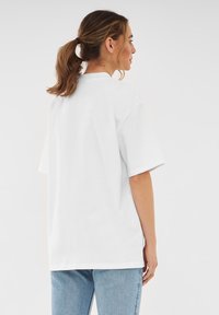 Vitt oversize t-skjorta med korta ärmar och klassisk rund halsringning, avbildad bakifrån, matchat med ljusblå jeans.