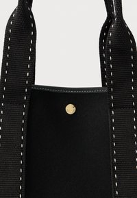Μαύρη τσάντα με υφή από ύφασμα και λευκές ραφές, με χρυσό κουμπί κλείσιμο και ανθεκτικά πλέ woven λουριά.