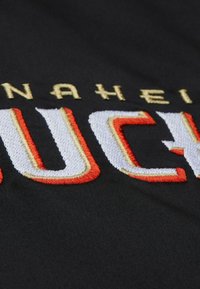 Schwarzer Stoff mit großem, gesticktem "ANAHEIM" in goldenen Buchstaben und "DUCKS" in Weiß und Orange, mit einer strukturierten, geschichteten Optik.