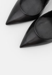 Ballerines noires en cuir, à bout pointu, avec une texture lisse et une découpe circulaire près de l'empeigne, présentant un design minimaliste et un éclat subtil.