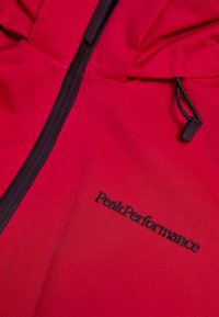 Rode softshell jack met zwarte rits en koord, voorzien van geborduurd zwart "Peak Performance" logo op de borst.