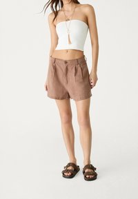 Shorts in lino marrone con pieghe frontali e chiusura con bottone, abbinati a un top bianco senza spalline e sandali marroni. Design semplice e texture leggera.
