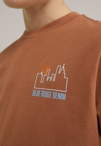 Rudas medvilninis džemperis su baltu miesto horizonto kontūru ir saulės grafika, pažymėta "BLUE RIDGE DENIM" mėlyna spalva.