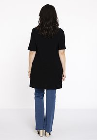Sort tunika med korte ærmer, der når til midt på låret, parret med lyseblå flared jeans og guld højhælede sko, set fra baghaven.