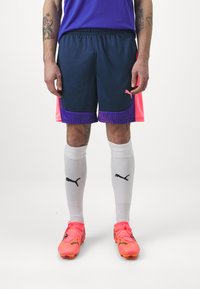 Marineblaue Sportshorts mit rosa Akzenten und lila Details, kombiniert mit weißen Socken mit schwarzen Logos und leuchtend pinken Fußballschuhen.