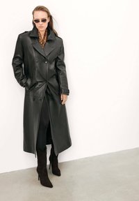 Abrigo de piel negro de doble botonadura con gran solapa y bolsillos laterales; llevado sobre leggings negros y botas de ante marrón oscuro de tacón alto.