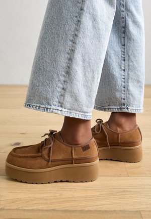 Person iført lyseblå vidde jeans og brune ruskindplatformsko med snørebånd, stående på et trægulv.