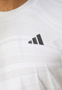 Valge sportlik T-särk tekstuuriga mustriga, millel on must Adidas'i logo rinnal. Kangas on sile ja kerge välimusega.