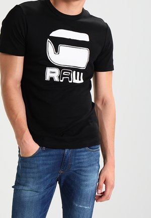 Mężczyzna w czarnej koszulce z dużym białym nadrukiem i napisem "RAW", ubrany w niebieskie dżinsy, stoi z jedną ręką w kieszeni.