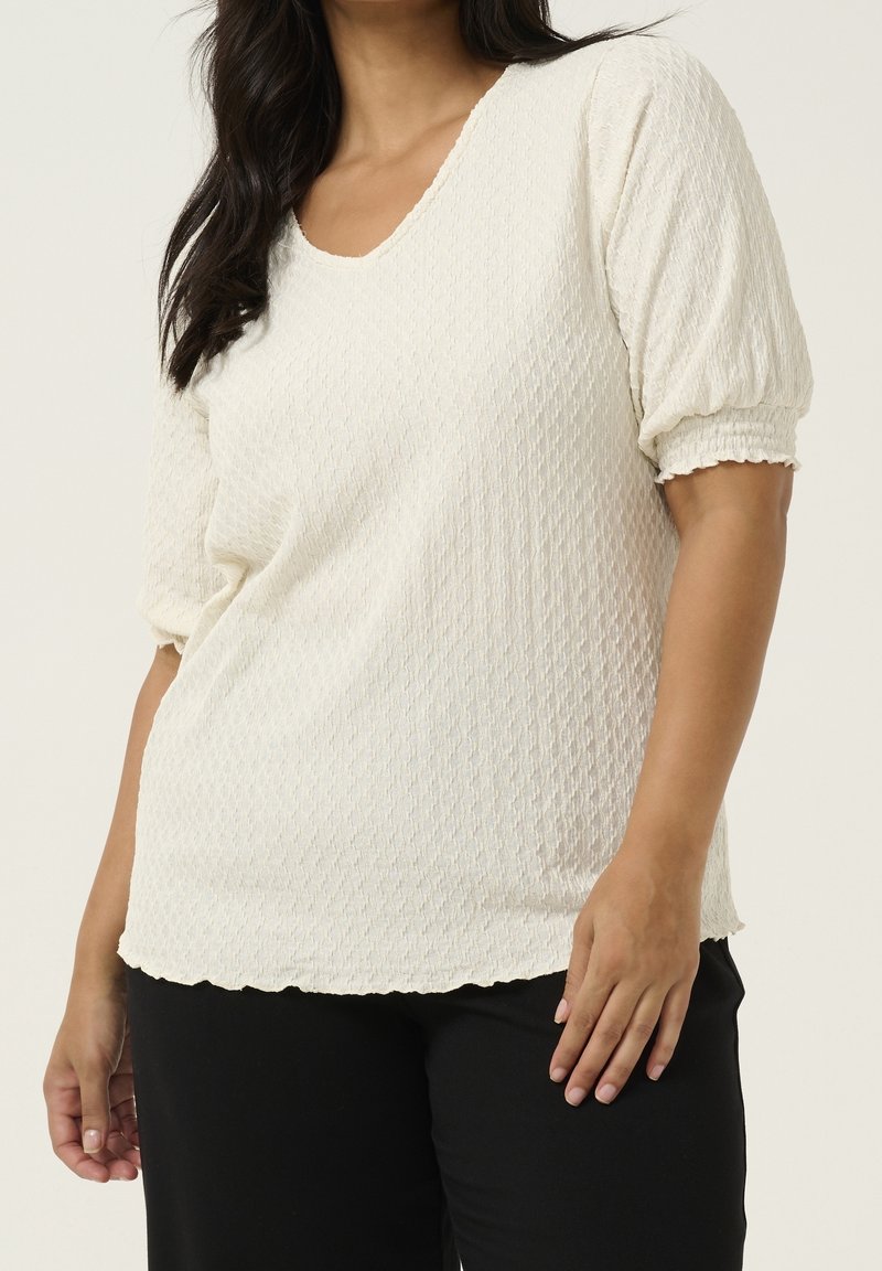 Haut blanc texturé avec un col en V et des manches courtes bouffantes. Il présente un ourlet festonné et un motif tissé pour un détail supplémentaire.