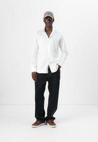 Camicia bianca a maniche lunghe, sbottonata, abbinata a jeans neri aderenti. Un cappellino beige e sneakers marroni con suole bianche completano il look.