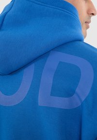 Haut à capuche bleu en tissu texturé, avec un grand logo délavé au dos. Le design comprend un capuchon à cordon de serrage et des poignets côtelés.