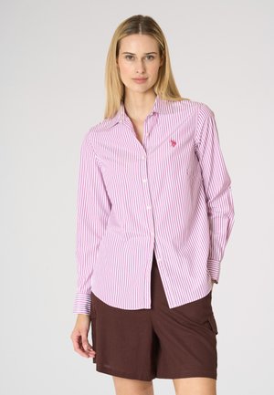 Donna in piedi che indossa una camicia a righe rosa e bianca a maniche lunghe con bottoni e pantaloncini marroni al ginocchio su uno sfondo semplice.