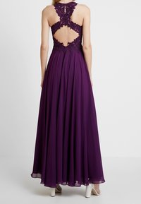 Fioletowa maxi sukienka z koronkowym gorsetem w kwiaty i otwartym plecami. Skórzany dół z lekkim, zwiewnym materiałem, wykończony warstwami tkaniny, oraz białe szpilki.