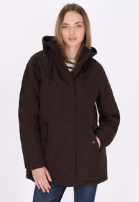 Giacca parka impermeabile marrone con cappuccio, dotata di chiusura frontale con zip, dettagli con coulisse e tasche laterali, indossata sopra una maglietta a righe.