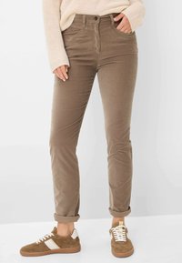 Braune Cordhosen mit tailliertem Design, umgeschlagenen Säumen, Fronttaschen und einem Knopfverschluss, kombiniert mit braunen und weißen Sneakers.