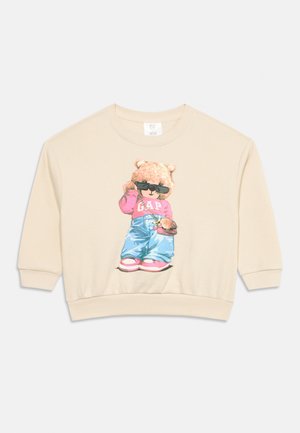 Sudadera beige con un oso que lleva gafas de sol, una sudadera rosa de GAP, pantalones azules, zapatos rosas y sostiene un bolso marrón.