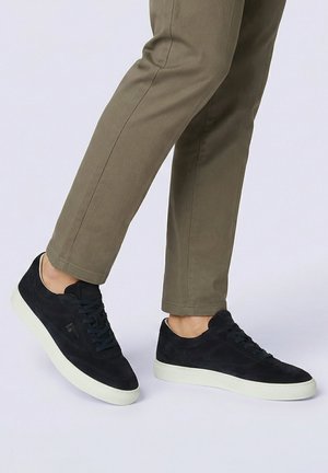 Person, die olivgrüne Hose und schwarze Wildledersneaker mit weißen Sohlen trägt, steht mit einem leicht angehobenen Fuß.