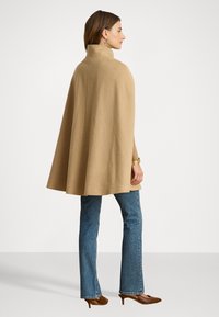 Μπεζ μάλλινο κάπες με ψηλό γιακά, ρευστός σχεδιασμός. Φορεμένο πάνω από ανοιχτό μπλε φαρδύ τζιν και καφέ ψηλοτάκουνα παπούτσια. Απλή, καθαρή σιλουέτα.