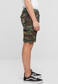 Karinės spalvos kargo šortai su žalia juosmens juosta, šoniniais kišenėmis ir kelių ilgio dizainu, derinami su juodais sportbačiais su baltais akcentais.