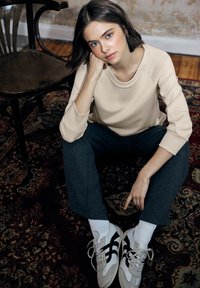 Top de manga larga beige, pantalones grises con rayas finas y deportivas blancas con detalles en negro. Sentado sobre una alfombra estampada junto a una silla de madera.