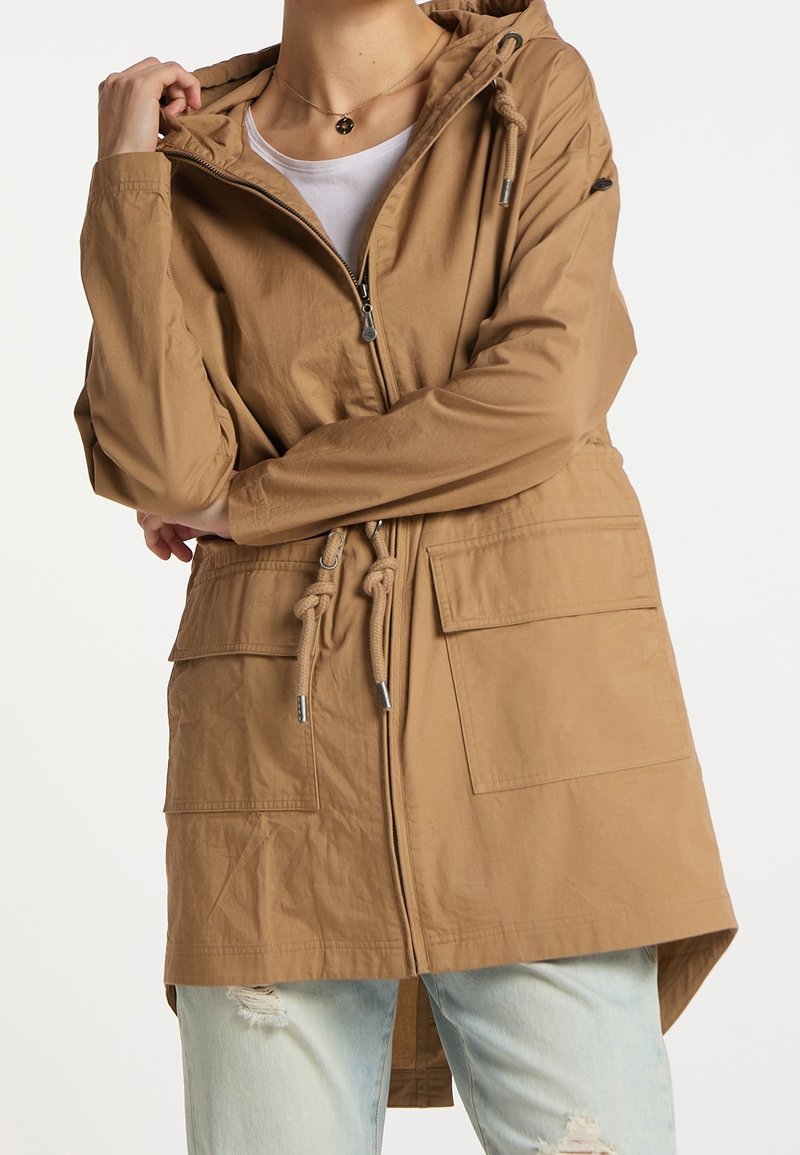 Personne portant une veste à capuche de couleur beige avec des poches avant, une fermeture éclair, une taille à cordon, associée à un jean bleu clair déchiré et une chemise blanche en dessous.