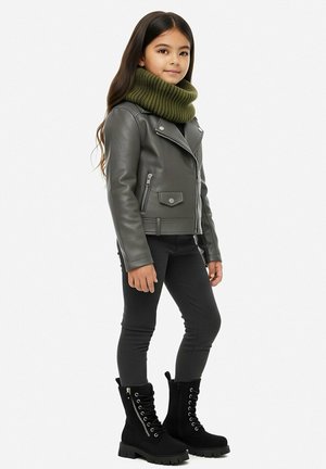 Junges Mädchen steht und trägt eine graue Lederjacke, schwarze Hose, schwarze Schnürstiefel und einen grünen Strickschal.
