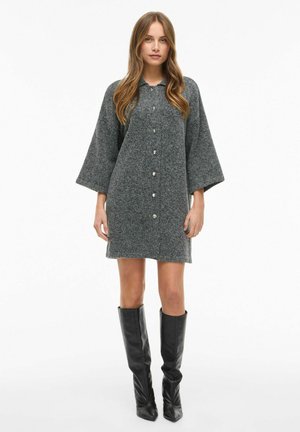 Šedé pletené šaty s knoflíkovým zapínáním vpředu, širokými rukávy a volným střihem, v kombinaci s vysokými černými koženými botami. Hladký povrch, bez vzorů.