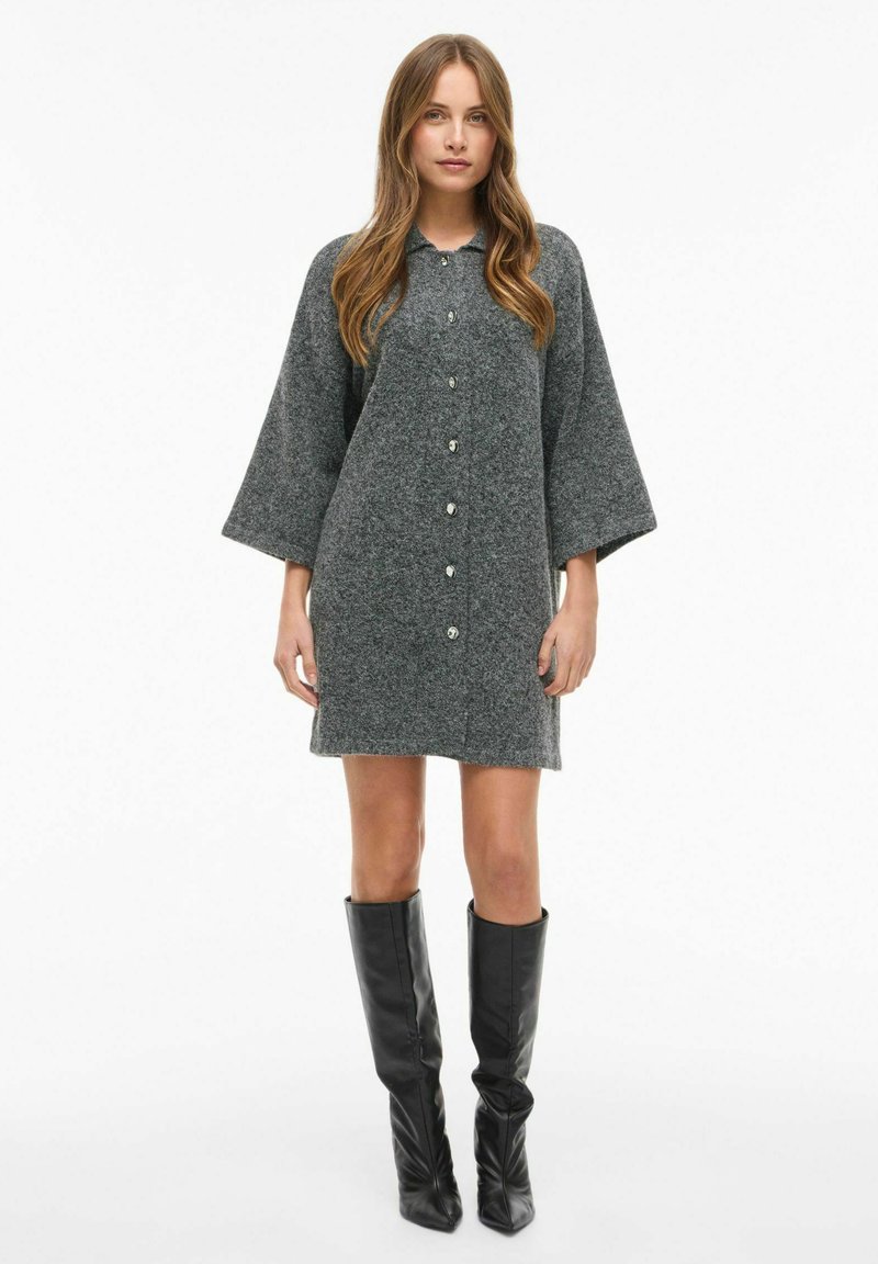 Pilka megzta suknelė su mygtukais priekyje, plačiomis rankovėmis ir laisvu siluetu, derinama su aukštomis juodomis odinėmis botais. Lygus audinys, be raštų.