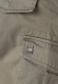 Närbild på en beige tygficka med sydda sömmar och en liten metallisk G-RAW-logotyp på fliken.