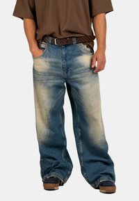 Person wearing loose, faded blue jeans with a brown woven belt, dark shoes, and a brown short-sleeve shirt, hands in pockets.