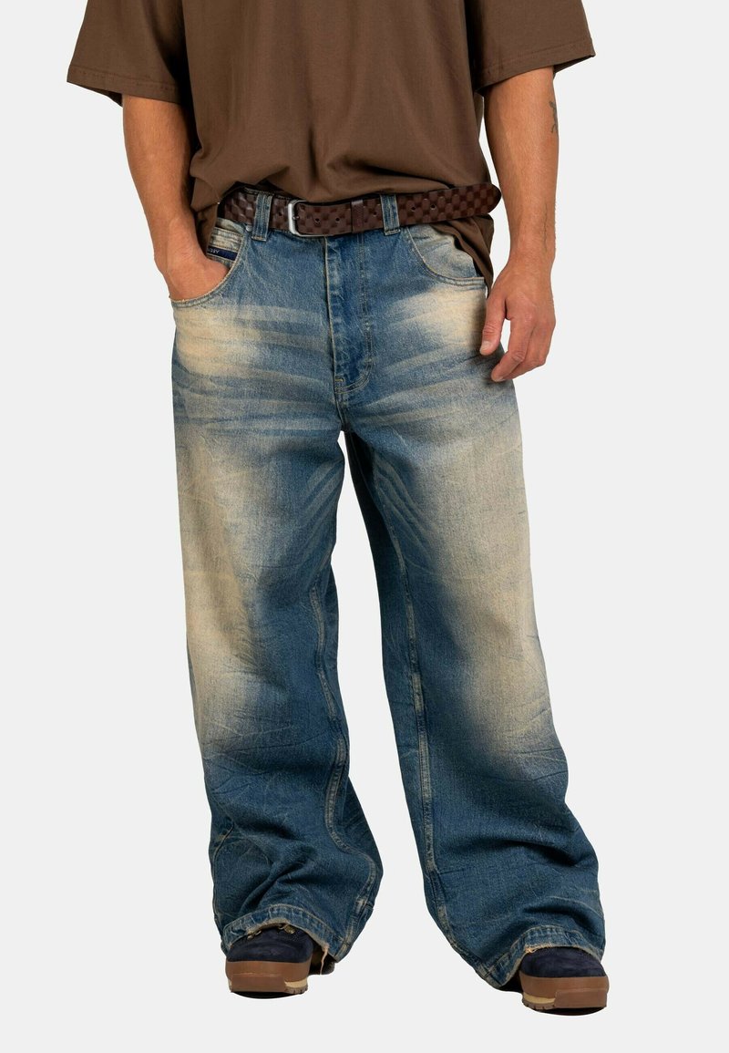 Person wearing loose, faded blue jeans with a brown woven belt, dark shoes, and a brown short-sleeve shirt, hands in pockets.