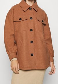 Brun uldblandingsjakke med krave og knaplukning fortil, der har to brystlommer og elastiske manchetter, præsenteret over en ribbet beige nederdel.