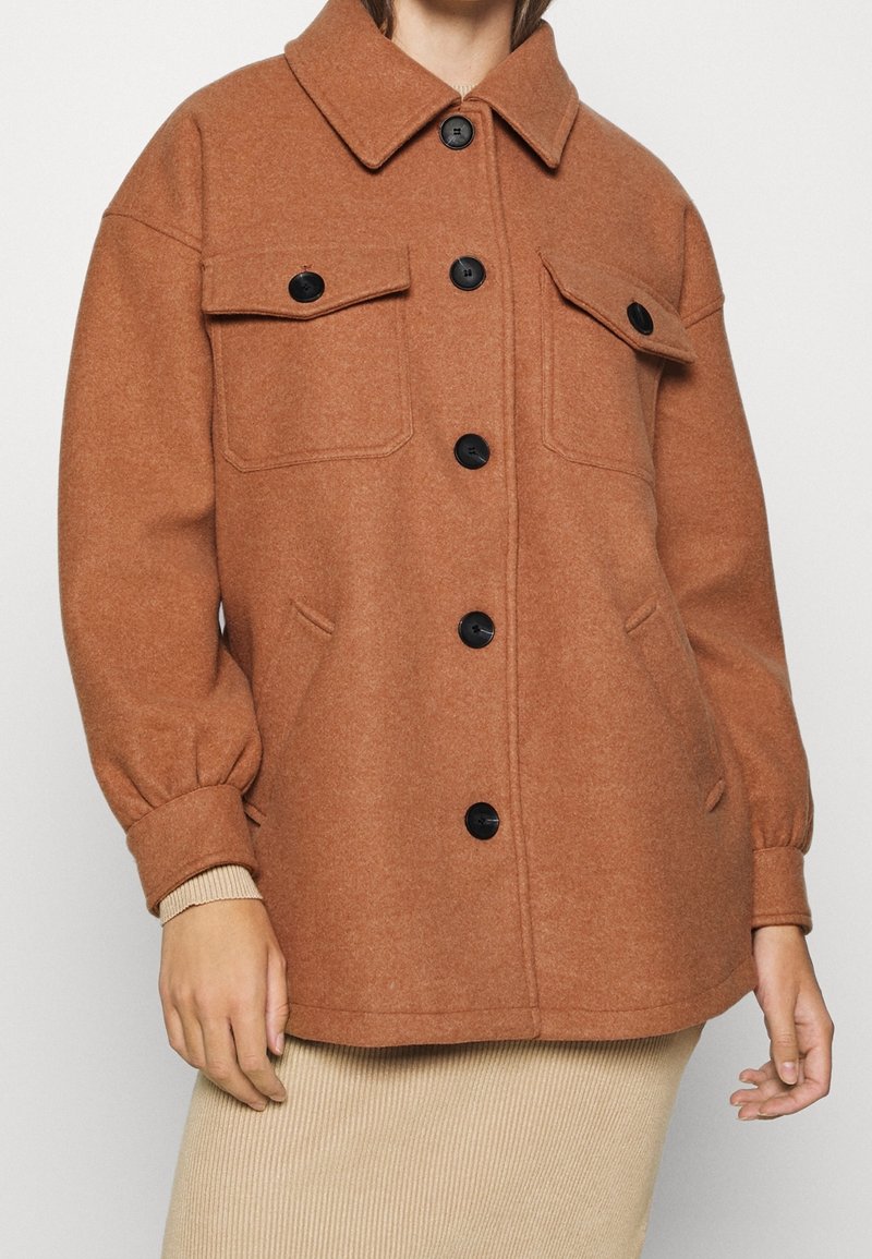 Brun uldblandingsjakke med krave og knaplukning fortil, der har to brystlommer og elastiske manchetter, præsenteret over en ribbet beige nederdel.