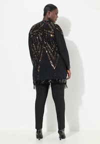 Chaqueta negra de manga larga con un patrón geométrico de lentejuelas, dobladillo con flecos y textura sedosa. Llevada sobre pantalones negros ajustados y zapatos de tacón alto.